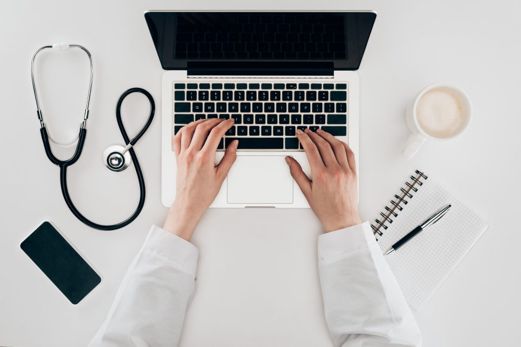 partial view of doctor at workplace with laptop, stethoscope, and cup of coffee